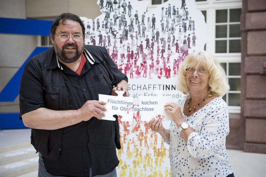 Foto von Peter Valtink und Zia Hüttinger beim Netzwerktreffen zu "Ausgezeichneter Ort 2016" in Berlin. Das Logo der Aktion steht im Hintergrund, davor links Peter Valtink, rechts Zia Hüttinger. Peter hält ein Puzzleteil auf dem "Flüchtlinge (zweite Zeile) für Ob" steht, Zia eins auf dem "kochen (zweite Zeile) dachlose" steht.