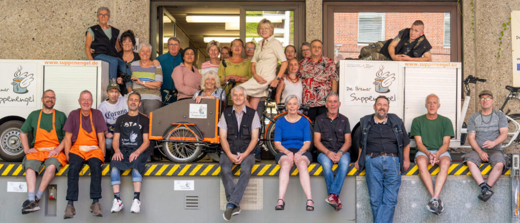 Foto vom Suppenengelteam. Vorne auf einer Kante sitzen acht Personen, Peter Valtik lehnt sich stehend an die Kante. Hinter den Personen stehen drei unserer Lastenfahrräder. Zwischen und auf den Fährradern sitzen und stehen weitere 17 Personen.