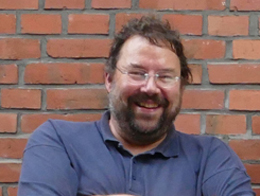 Foto von Peter Valtink, unserem Geschäftsführer. In Hintergrund sind die Steine einer Backsteinmauer zu sehen. Peter lacht frontal in die Kamera. Er hat kurze, dunkelbraune Haare, eine randlose Brille und einen dunkelbraunen Vollbart, der seitlich grau ist. Er trägt ein dunkelblaues Polohemd.