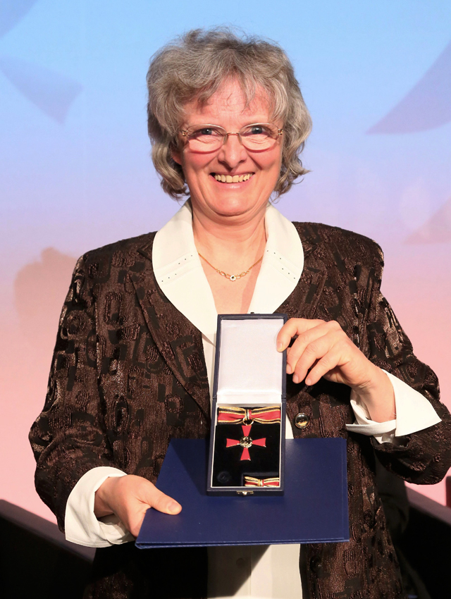 Foto der Gründerin der Bremer Suppenengel Zia Gabriele Hüttinger. Im Hintergrund ist der Ausschnitt einer blau und rosa beleuchteten Wand zu sehen. Im Vordergrund steht Frau Hüttinger. Sie lächelt und hält die geöffnete Schachtel mit dem Bundesverdienstkreuz in der Hand. Das rote Kreuz liegt auf einer schwarzen, samtigen Unterlage und hat oben eine silberne Aufhängung, durch die ein Band läuft. Das Band hat einen breiten, roten Streifen in der Mitte. Darüber und darunter jeweils zwei goldene und einen schwarzen dünnen Streifen. Frau Hüttinger hat kinnlange, gewellte graue Haare und eine Brille. Sie trägt eine braune, etwas glänzende, elegante Jacke, darunter eine weiße Bluse mit großen Aufschlägen an Kragen und Ärmeln.