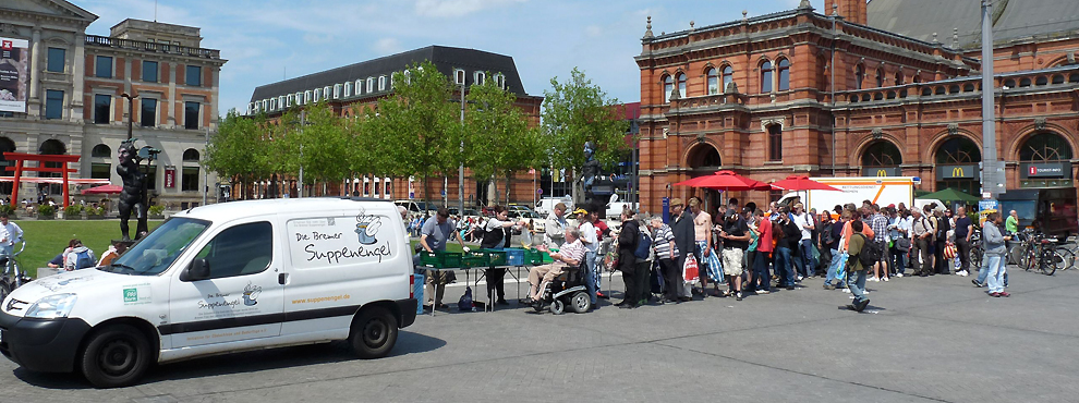 Foto von der Ausgabe von Lebensmitteln durch die Suppenengel am Bremer Hauptbahnhof. Im Hintergrund sind der Hauptbahnhof und das Überseemuseum zu sehen, davor ganz links ein weißes Auto mit dem Logo der Bremer Suppenengel. Rechts vom Auto stehen Tische mit grünen Kisten darauf, dahinter Menschen, die den Inhalt der Kisten verteilen. Entlang der Tische und darüber hinaus hat sich eine Schlange von anstehenden Menschen gebildet. Die erste Person in der Schlange sitzt in einem Elektro-Rollstuhl.