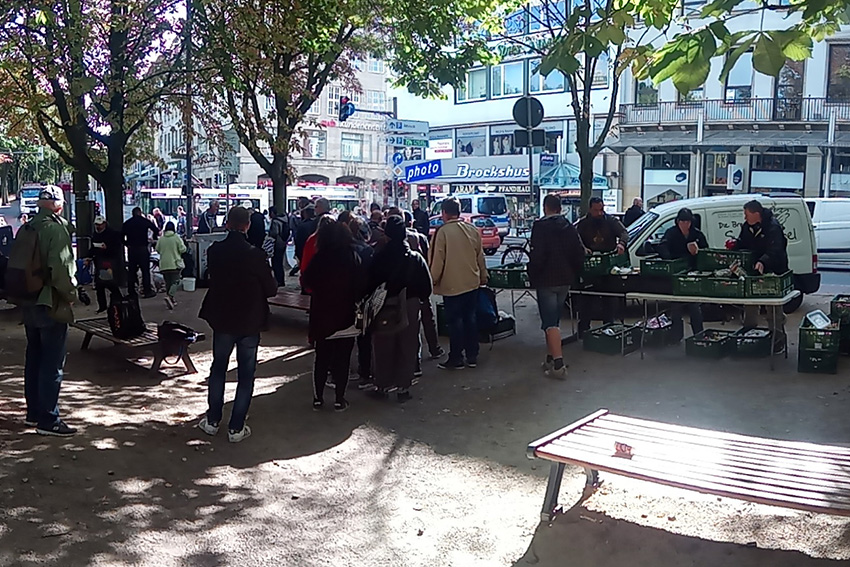 Foto von der Essensausgabe am Kaisen-Denkmal. Unter den Bäumen der Wallanlagen sind Tische mit grünen Boxen darauf aufgebaut. Dahinter und davor stehen Menschen. Im Hintergrund ein Suppenengel-Auto. Ganz hinten ist Photo Brockshus und eine Straßenbahn zu sehen.