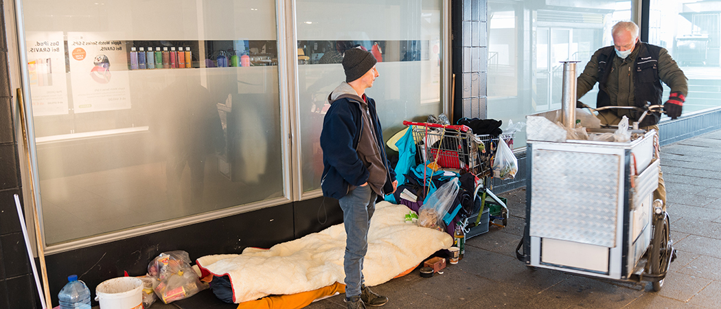 Foto vor einer Schaufensterfront mit milchig abgeklebten Scheiben. Links im Bild ist die Schlafstelle und die Habseligkeiten der Person die davor steht. Die Person trägt eine dunkle Mütze, eine dunkelblaue Jacke, einen dunkelbraunen Pullover und eine graue Hose. Sie blickt zu einem Mann, der von rechts ins Bild kommt. Der Mann fährt eines unserer Lastenräder. Er trägt einen Mundschutz, eine dunkle Jacke und dicke Handschuhe.