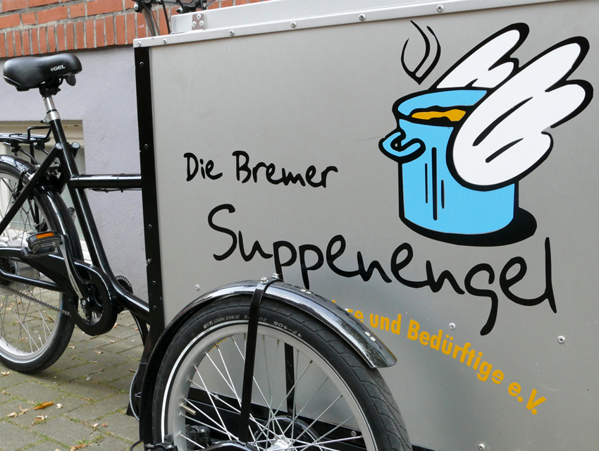 Foto von einem unserer Lastenfahrräder. Eine Nahaufnahme eines Fahrrads von schräg vorne. Rechts und groß im Bild ist die silberne Transportbox des Fahrrads mit unserem Logo darauf. Links ist der schwarze Rahmen und Sattel des Rades zu sehen.