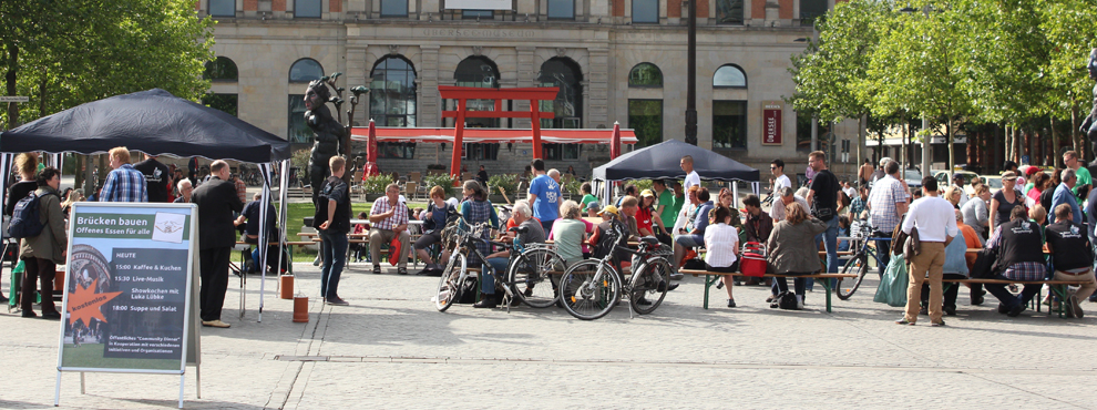 Foto von der Aktion "Brücken bauen - Offenes Essen für alle". Im Hintergrund ist das Überseemuseum zu sehen, davor steht eine rote, asiatische Konstruktion. Im Vordergrund stehen zwei schwarze Pavillions und mehrere Biertischgarnituren, an denen Menschen sitzen. Links im Bild ist eine Hinweistafel für unsere Aktion.