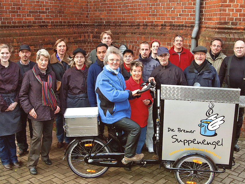 Foto von Menschen, die bei uns im Verein aktiv sind. Im Hintergrund ist eine Backsteinmauer. Davor stehen 16 Menschen, vor den Menschen steht eins unserer Lastenfahrräder mit einer kleinen silbernen Transportbox hinten und einer großen silbernen Transportbox vorne. Auf der vorderen Box ist unser Logo. Auf dem Fahrrad sitzt unsere Gründerin Zia Hüttinger.