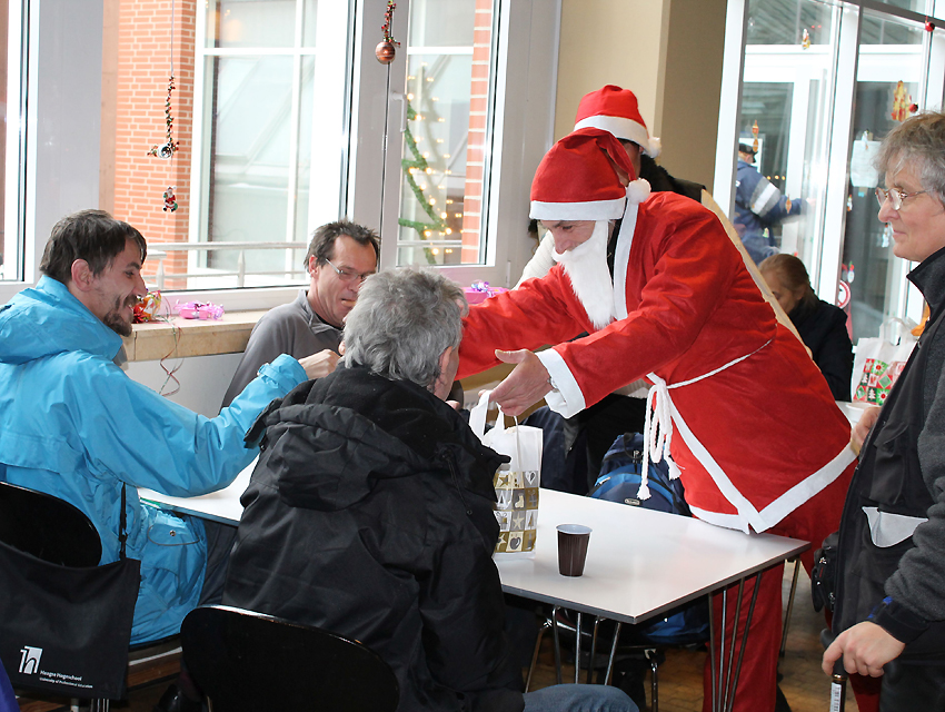 Foto von der Weihnachtsaktion. Im Hintergrund ein Fenster mit ein bisschen weihnachtlichem Schmuck. Im Vordergrund steht ein weißer Tisch, an dem drei Männer sitzen. Ein als Nikolaus verkleideter Mann steht am Tisch und stellt einen weihnachtlich dekorierten Beutel auf den Tisch. Ganz rechts im Bild ist eine Person im Profil zu sehen.