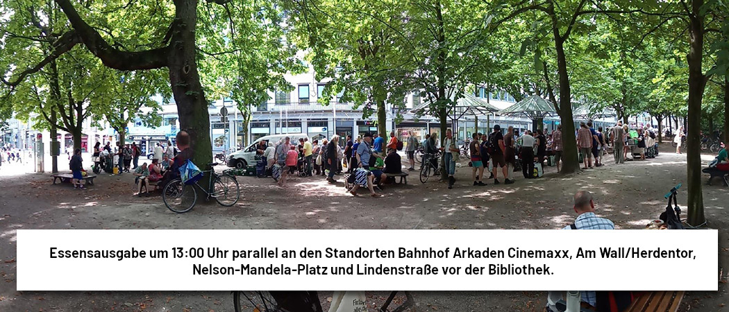 Foto von der Essensausgabe Am Wall/Herdentor. Es sind viele grüne Bäume zu sehen, dazwischen stehen sehr viele Menschen. Auf dem Bild der Text: Essensausgabe um 13:00 Uhr parallel an den Standorten Bahnhof Arkaden Cinemaxx, Am Wall/Herdentor, Nelson-Mandela-Platz und Lindenstraße vor der Bibliothek.