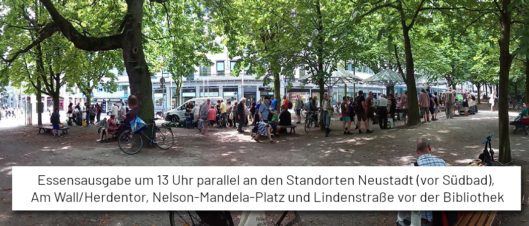 Foto von der Essensausgabe Am Wall/Herdentor. Es sind viele grüne Bäume zu sehen, dazwischen stehen sehr viele Menschen. Darunter steht "Essensausgabe um 13 Uhr parallel an den Standorten Neustadt (vor Südbad), Am Wall/Herdentor, Nelson-Mandela-Platz und Lindenstraße vor der Bibliothek".